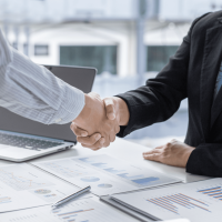 Two people shaking hands over a table with papers and a laptop.